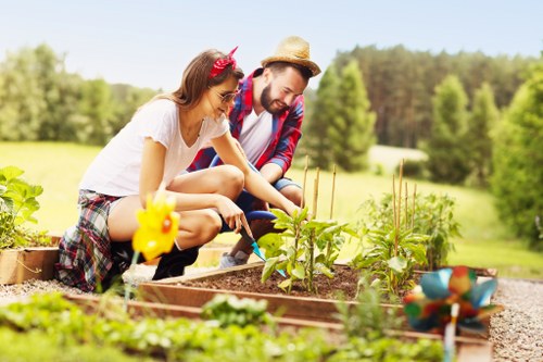 In a bright and sunny outdoor garden setting with lush greenery and trees in the background, a woman and a man are engaged in gardening activities. The woman, wearing a white top and plaid shorts, is crouched down tending to a small garden bed with various young plants. She has a red headband and appears focused on her task. The man, dressed in a plaid shirt, white T-shirt, and a straw hat, is also crouched nearby, observing or assisting her. The garden bed is bordered with wooden planks and contains soil and emerging green plants, some supported with small stakes. In the foreground, adjacent to the garden bed, there is a small rake and a decorative garden ornament resembling a yellow duck, while the background features a neatly maintained lawn and mature trees, creating a peaceful, natural outdoor environment. The scene suggests a local Wanstead garden cared for by specialists such as Landscapers Wanstead, highlighting healthy plant growth and active outdoor gardening amid pleasant weather conditions.