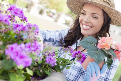 A woman wearing a straw hat and gardening gloves is engaged in planting or tending to colorful flowering plants in a well-maintained outdoor garden space. In the foreground, bright purple and pink flowers are prominently visible, with green foliage adding to the vibrant scene. The garden features a lush, green lawn area and a neatly arranged flower bed, bordered by a stone or brick edging. The background shows a slightly blurred view of trees and possibly a garden path or patio, suggesting a residential setting in Wanstead. The scene is brightly lit, indicating a sunny day with natural light enhancing the cheerful atmosphere. This image exemplifies garden maintenance and planting activities typical of outdoor landscaping and gardening services, emphasizing the care and attention given to creating aesthetically pleasing garden environments.