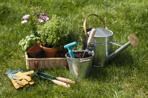 A neatly maintained garden with a lush, green lawn displaying dense grass and a variety of garden tools and potted plants arranged on the grass. In the foreground, there are yellow gardening gloves, a pair of small hand secateurs, and a garden fork placed beside a metal watering can with a wooden handle. To the left, a rectangular wicker basket contains several potted plants, including flowering and foliage varieties, with some plants having purple and green leaves. Behind the basket, additional plant pots of various sizes are visible, contributing to a vibrant, cultivated outdoor space. The scene suggests early morning or late afternoon light, casting soft shadows on the garden. This environment exemplifies typical outdoor gardening tasks, such as planting, watering, and tending to flower beds, reflecting professional landscaping and gardening services available in Wanstead, London, as offered by Landscapers Wanstead, who specialise in creating well-kept and visually appealing outdoor spaces.