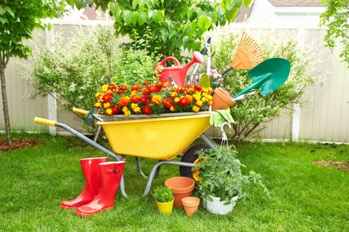 A bright, colorful garden scene in a residential backyard with a vibrant green lawn bordered by a white picket fence. Central to the image is a yellow garden wheelbarrow filled with red and orange flowers, positioned on the grass. Surrounding the wheelbarrow are various gardening tools and accessories, including red rubber boots placed on the ground nearby, a small yellow pot, and three terracotta plant pots of different sizes. Behind the wheelbarrow, there is a red watering can resting beside two shovels with orange and teal blades, both leaning against the fence. A white plant pot with a green leafy shrub is situated to the right, and additional greenery, including trees and shrubs, provides a lush background, creating a natural outdoor environment. The scene appears to be set on a bright, partly cloudy day, suitable for garden maintenance, with a focus on gardening activities typical of landscaping services in Wanstead or nearby areas. The composition highlights the tools, plants, and vibrant outdoor setting representative of professional gardening and landscaping work available from Landscapers Wanstead.