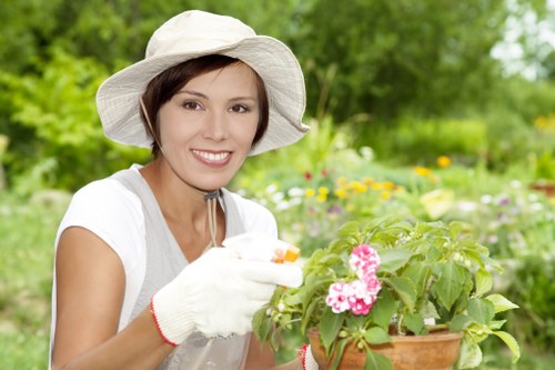 A woman with short brown hair, wearing a wide-brimmed white gardening hat and white gardening gloves, is smiling while tending to a potted flowering plant with pink blossoms. She is outdoors in a lush garden setting with bright green foliage, including bushes and trees, in the background. The garden features a well-maintained lawn with vibrant green grass, bordered by flower beds and shrubbery, indicative of a landscaped outdoor space typical for residential gardens in Wanstead. The scene suggests a mild, sunny day, emphasizing natural light illuminating the garden's varied textures and colours. This image reflects professional gardening and landscaping activities, highlighting plant care within a garden environment that could be serviced by providers like Landscapers Wanstead, especially in the Wanstead area or nearby postcode regions. The overall composition showcases attention to detail in plant maintenance, ideal for enhancing outdoor spaces through garden and lawn care services in London Borough of Redbridge or surrounding areas.
