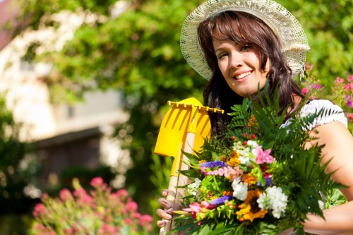 A woman with wavy brown hair and a broad smile, wearing a wide-brimmed straw hat and a sleeveless white top, standing outdoors in a well-maintained garden. She is holding a yellow garden rake in her right hand and a large, vibrant bouquet of various flowers and green foliage in her left arm. The garden features a lush, green lawn in the foreground, bordered by flower beds with colorful blooms, and dense trees and shrubs in the background providing a natural, shaded environment. The scene is illuminated by natural daylight, suggesting a clear, sunny day, and the overall setting reflects a tidy, landscaped outdoor space suitable for gardening services in Wanstead or nearby areas. The image emphasizes outdoor maintenance and gardening activities, aligning with landscaping and lawn care themes typical for local gardens in the London borough.