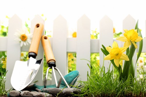 The image depicts a section of a well-maintained garden with a white picket fence in the background, possibly part of a front or back garden landscape typical in Wanstead. In the foreground, there is a patch of lush green grass with dense, healthy growth and a few yellow daffodils in bloom, adding vibrant natural colour to the scene. Adjacent to the grass, a small flower bed is defined by soil and contains blooming yellow daffodils with elongated green leaves. Leaning against the fence are two gardening tools: a small hand trowel with a wooden handle and a garden fork with a teal-colored handle, suggesting active garden maintenance. The environment appears bright and sunny, indicative of good weather conditions that support outdoor gardening activities. This scene emphasizes typical garden elements such as lawns, flower beds, and gardening tools, aligning with landscaping and garden maintenance services offered by companies like Landscapers Wanstead, serving the local Wanstead area within E11 postcode. The overall composition showcases a neat, natural outdoor space ready for further gardening or decking repair work, contributing to the aesthetic appeal of residential outdoor environments in this region.