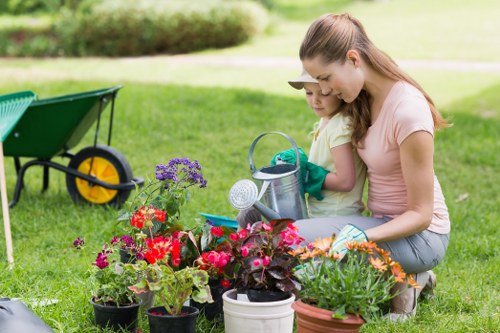 In a well-maintained outdoor garden area, two children are engaged in planting activities amidst a variety of flowering plants and potted greenery. The scene features a lush, green lawn with neatly trimmed grass, and a collection of vibrant flowers including red, purple, and orange blossoms arranged in pots and garden beds. A garden watering can and gardening gloves are visible, indicating active gardening work. In the background, there is a green wheelbarrow with black handles, resting on the grass, and further out, a hedge line or shrubbery borders the garden space, providing a natural backdrop. Soft natural light suggests a clear, mild day, creating an inviting environment typical of outdoor gardening in Wanstead or nearby areas. The children appear focused on their planting tasks, supported by the landscaping and gardening services offered by Landscapers Wanstead, who specialise in creating beautiful, functional garden landscapes in the local area. Overall, the scene captures a peaceful, productive moment of outdoor planting in a suburban garden, emphasizing the beauty of garden elements and the importance of professional gardening care in the UK.