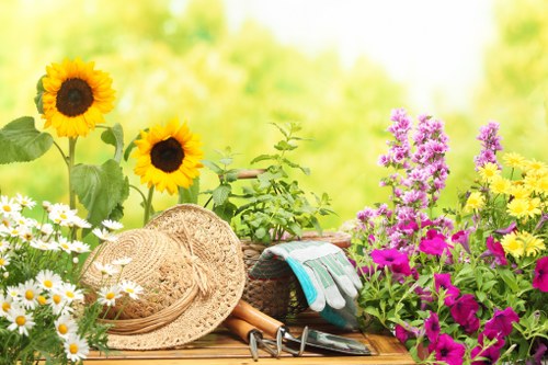 A vibrant garden scene featuring a variety of flowering plants and greenery, including tall sunflowers with large yellow petals and dark centers, clusters of purple and pink flowers, and white daisies with yellow centers, all arranged along a wooden garden table or bench. Resting on the table are gardening tools such as a strimmer, a small pair of pruning shears, gardening gloves, and a straw hat, suggesting outdoor gardening activity. The background is filled with bright, natural daylight, highlighting the lush green foliage and creating a warm, inviting atmosphere in a neatly maintained outdoor space. This setting reflects typical features of a well-kept, landscaped garden in Wanstead, suitable for gardening, lawn care, and outdoor maintenance services by Landscapers Wanstead, incorporating natural textures like soil and plant foliage, with vibrant colour variations that enhance the visual appeal of a residential garden environment.