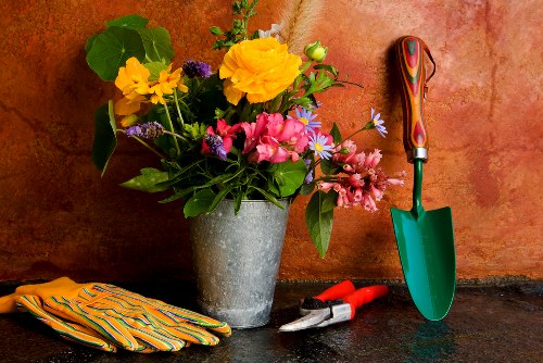 The image displays a collection of gardening tools and decorative elements arranged on a dark stone surface, set against a warm, orange-toned wall. In the foreground, there is a metal bucket filled with a vibrant assortment of fresh flowers, including yellow, pink, and purple blossoms with green foliage, creating a lively display. To the right, a small green-handled trowel with a colorful wooden grip is planted into the surface, indicating outdoor gardening activity. Nearby, red-handled pruning shears rest alongside a pair of yellow gardening gloves with black accents, all suggesting readiness for plant maintenance. The outdoor environment appears well-maintained and suitable for landscaping work typical of Wanstead, reinforcing the relevance of local gardening services. The scene captures a moment of preparation or decoration for a garden or outdoor space, with natural lighting emphasizing the bright colors and textures of the flowers, tools, and accessories, suitable for illustrating gardening and landscaping activities associated with Landscapers Wanstead on their Recycling and Sustainability page.