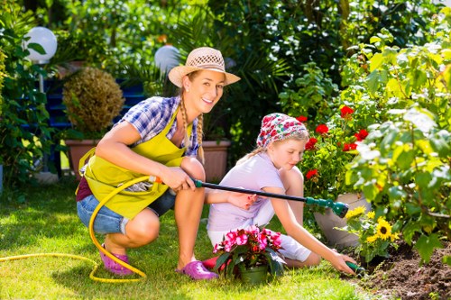 In a lush, well-maintained garden with vibrant green grass and a variety of flowering plants and shrubs, a woman and a young girl are engaged in gardening activities. The woman, dressed casually in a yellow apron over a checked shirt and wearing a straw sun hat, is smiling as she holds a garden hose, indicating she is watering the plants. The girl, wearing a floral sun hat and a light-colored dress, is focused on planting or tending to flowers in a flower bed bordered by a small, neat edge. Surrounding them are flowering plants with red, yellow, and pink blossoms, set against a backdrop of dense trees and hedges, providing privacy and a natural setting typical of gardens in Wanstead. The scene appears to be during daytime with bright natural light, highlighting the healthy, lush foliage and colorful blooms, reflecting professional gardening work by Landscapers Wanstead in a community garden or backyard setting aimed at outdoor maintenance and aesthetic enhancement.