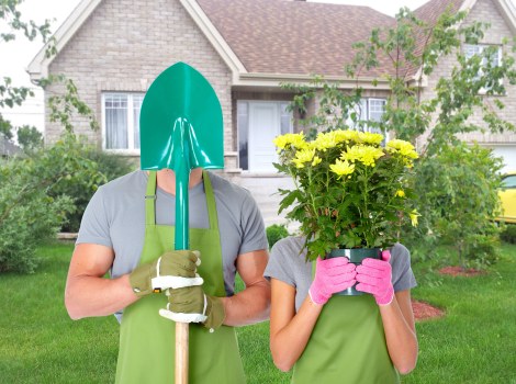 A garden scene showing two individuals in the front yard of a suburban property, with a background of a brick house featuring white-framed windows and a pitched roof. The person on the left, wearing a grey t-shirt, green gardening gloves, and a large green sun hat, holds a green-handled spade vertically, covering their face with the hat's brim. The person on the right, dressed in a grey top and pink gardening gloves, is holding a large, lush bouquet of yellow flowers with green foliage in front of their face, obscuring their identity. The garden includes a well-maintained lawn with dense, green grass, bordered by shrubs and small trees, and a designated flower bed area in the background. The overall environment appears bright and outdoors, reflecting seasonal gardening activities in Wanstead, with the scene captured on a clear day, emphasizing outdoor landscaping and planting work.