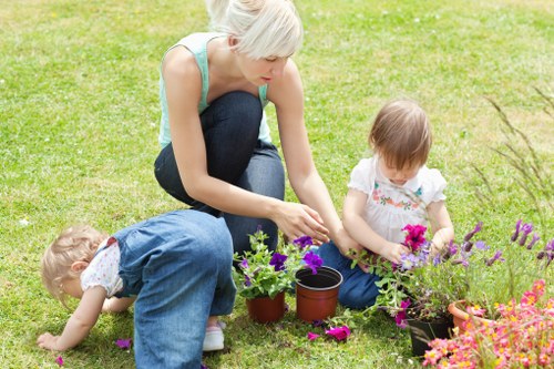 A woman with short blonde hair, wearing a sleeveless light blue top and dark jeans, is kneeling on a well-maintained grassy lawn in Wanstead, engaging in gardening activities alongside two young children. The children, both with light brown hair, are also kneeling on the grass; one is dressed in a floral top and denim shorts, while the other is in a white dress with colorful patterns. They are tending to flower pots filled with vibrant, blooming flowers in shades of purple, pink, and green, with some flowers scattered on the grass around them. In the background, the lush lawn extends, bordered by a variety of shrubs and possibly small trees, indicating a landscaped garden space suitable for outdoor family activities. The scene takes place during daytime with natural light, reflecting a peaceful outdoor environment that emphasizes well-kept lawns, garden plants, and landscaping features typical of Wanstead's residential gardens, aligning with gardening services offered professionally by Landscapers Wanstead.