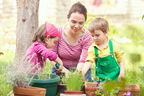 A woman with dark hair tied back, dressed in a pink and white striped top, is outdoors in a well-maintained garden during daylight, tending to a variety of potted plants and flowers placed on a garden table, with children beside her engaged in gardening activities; behind her, a sturdy tree trunk and lush green grass with small flowering plants are visible, suggesting an organized and cultivated outdoor space within a residential garden, which may be part of landscaping or gardening services in Wanstead, Essex, as indicated by the context. The scene captures the peaceful, natural setting of a landscaped yard, ideal for gardening activities, with natural lighting highlighting the vibrant colours of the plants and the casual, engaging atmosphere of outdoor plant care, typical in local garden maintenance and flower bed creation.