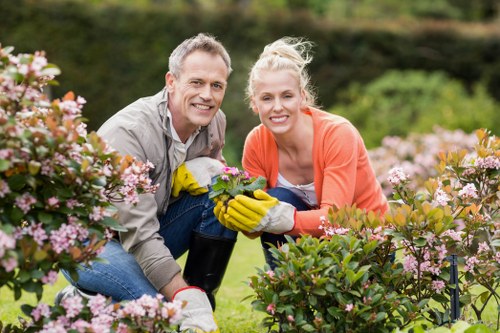 A man and a woman are crouching in a well-maintained garden, surrounded by pink flowering shrubs and green foliage. The man, dressed in a light grey shirt, and the woman, wearing an orange cardigan, are both wearing yellow gardening gloves and smiling at the camera while holding a small potted plant together. The garden features lush, dense grass and a variety of plants arranged in borders, with a backdrop of trees and a hedge. The scene depicts outdoor gardening activity in a landscaped yard, reflecting professional gardening and landscaping services by Landscapers Wanstead, with natural lighting and a clear, mild weather condition typical of a suburban garden in Wanstead, London. The overall setting highlights a healthy, vibrant outdoor space suitable for lawn care, pruning, and planting tasks.